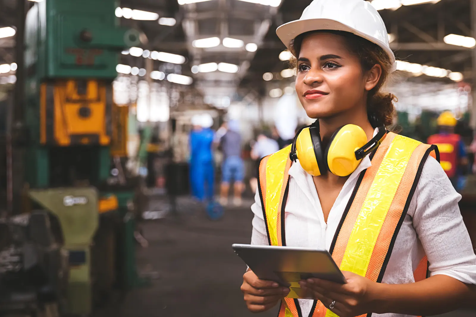Mujer con casco blanco, chaleco reflectivo y protectores auditivos sosteniendo una tableta en una planta industrial